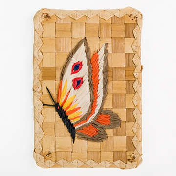 A woven rectangular panel made of light and dark natural fibers featuring a colorful butterfly crafted from dyed straw. The butterfly has orange, red, black, and cream highlights and is centered on the woven surface.