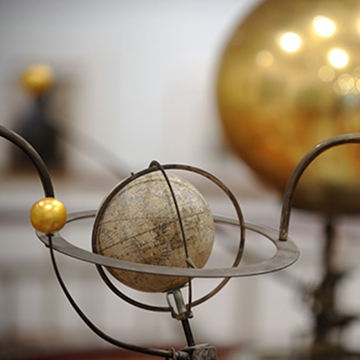 A close-up of an antique orrery-style scientific model featuring a small earth-like sphere encircled by metal rings, with a smaller yellow sphere attached to an arm. A large golden sphere appears blurred in the background.