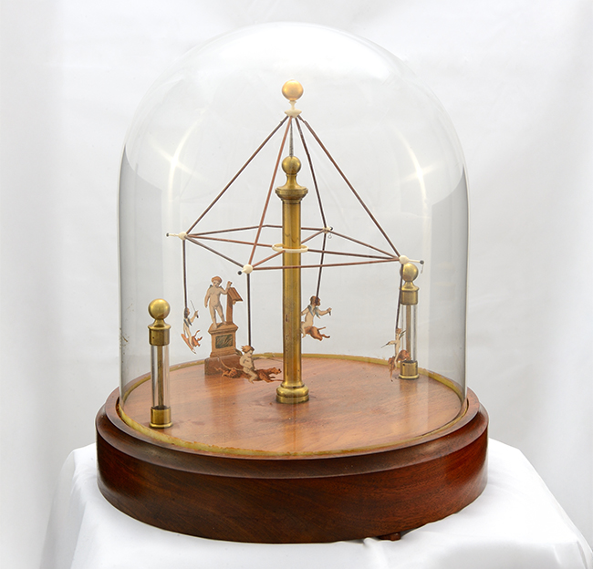A 19th-century perpetual motion demonstration device displayed under a glass dome. The apparatus features a central brass column supporting a geometric frame with suspended carved figures that appear to ride animals, all set on a polished wooden base.
