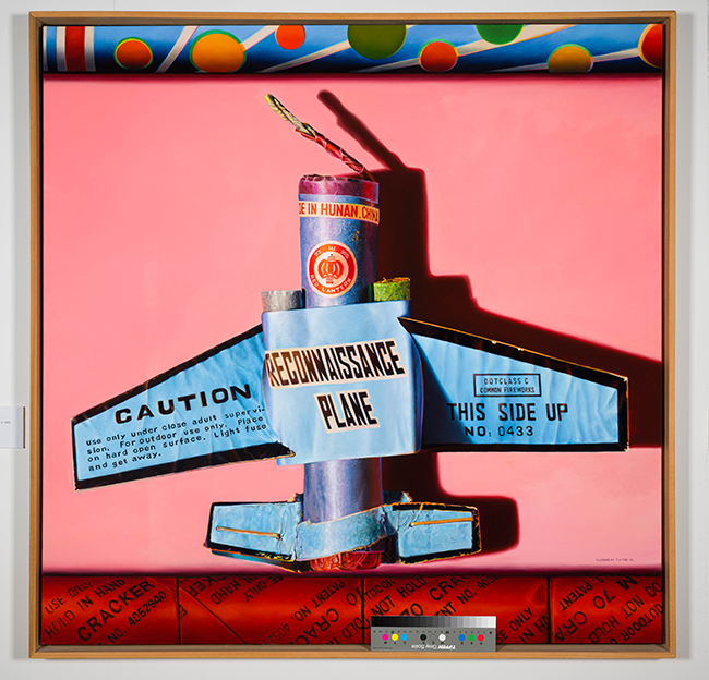A brightly colored painting depicting a blue cardboard “reconnaissance plane” constructed from firework packaging, set against a solid pink background. The plane’s body and wings feature caution labels and printed text, with a fuse emerging from the top. A rolled, patterned border in blue, orange, and green appears at the top of the composition.