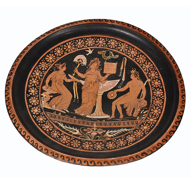 A circular ancient Greek red-figure plate decorated with three finely detailed figures. Two standing women in flowing garments flank a seated nude male figure, all positioned around a large central vessel. Floral and geometric patterns form an ornate border around the scene.