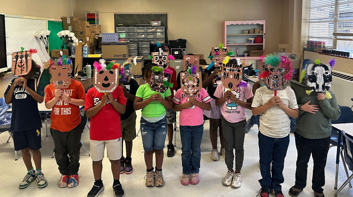 A group of elementary students stand in a classroom holding up colorful handmade masks decorated with feathers, beads, and paper cutouts. Art supplies and storage bins are visible in the background.
