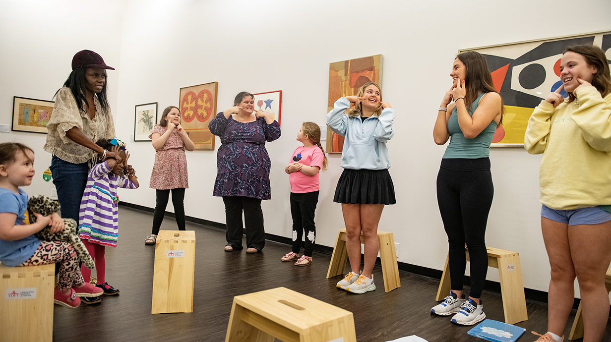 Participants in the Building Brains program at the University Museum stand in a circle smiling and following along with a group activity led by UM School of Education facilitators. The free, drop-in morning workshop welcomes adults and their children for story, music, art, and sensory play on the museum grounds.