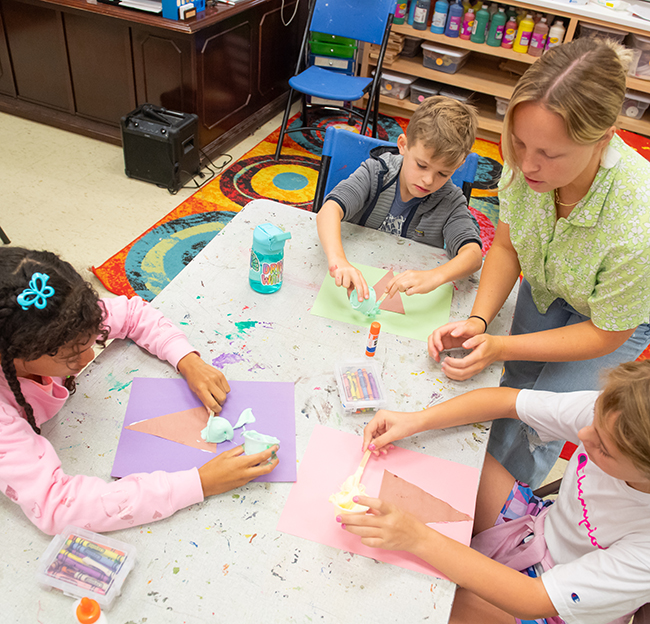 Children create art with paper and modeling materials at a classroom table, with an adult assisting one of the students.