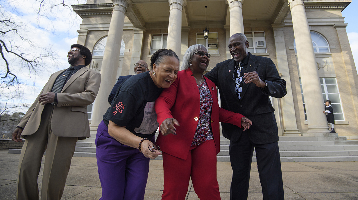 Two men and two women share a hearty laugh outside a tan stone building with large columns.