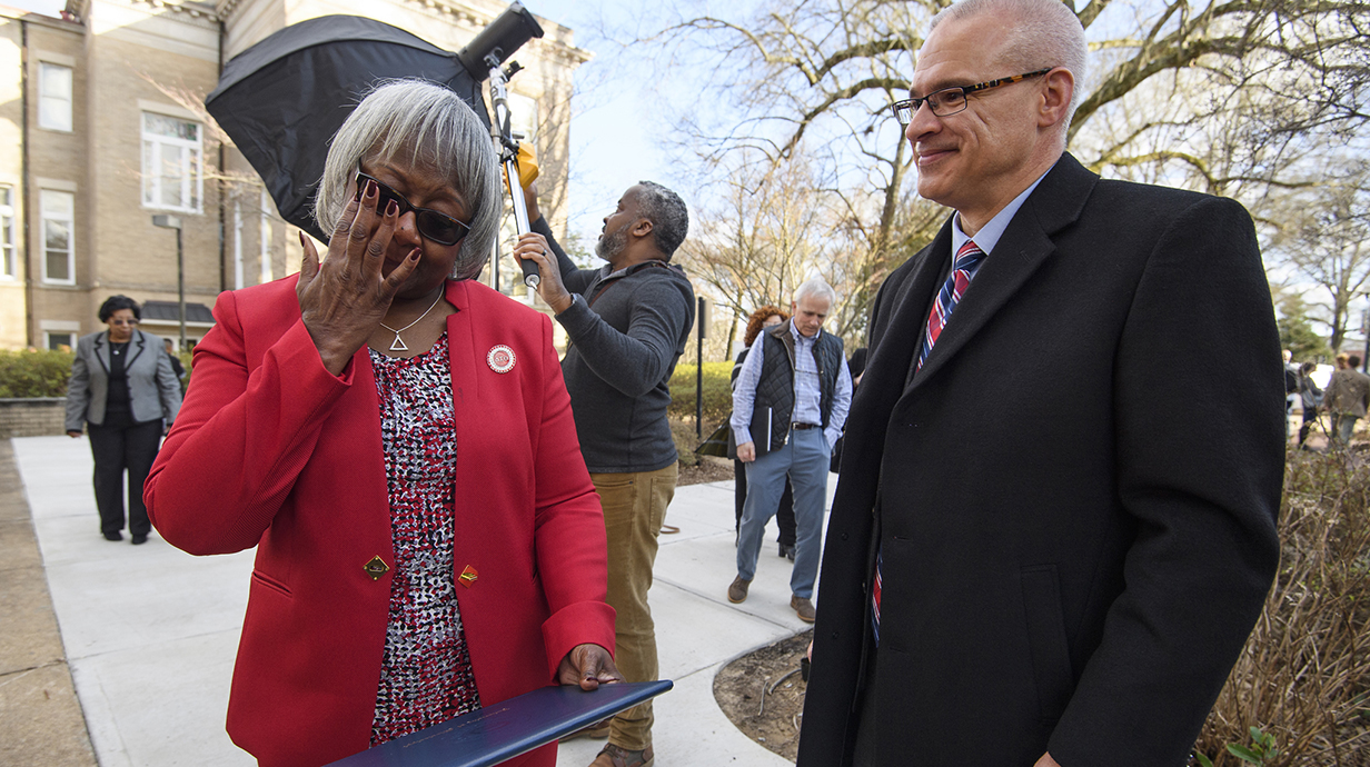A woman wearing a red pantsuit wipes her eye while talking to a man wearing a dark suit outside a large stone building.