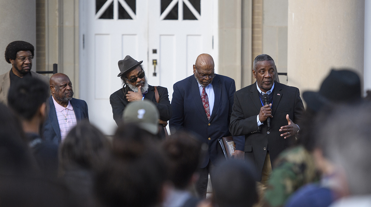 A man speaks into a microphone as a crowd watches on the frpnt porce of a large stone building.
