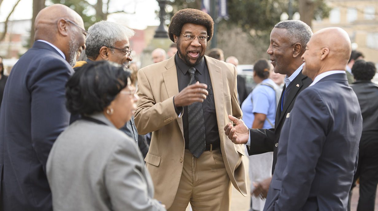 A man wearing a tan suit gestures as he talks with a group of people standing outdoors.