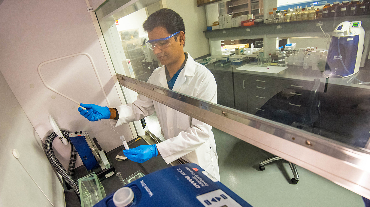A man wearing a white lab coat and blue gloves uses a pipette to transfer liquid into a container underneath a vented lab hood.