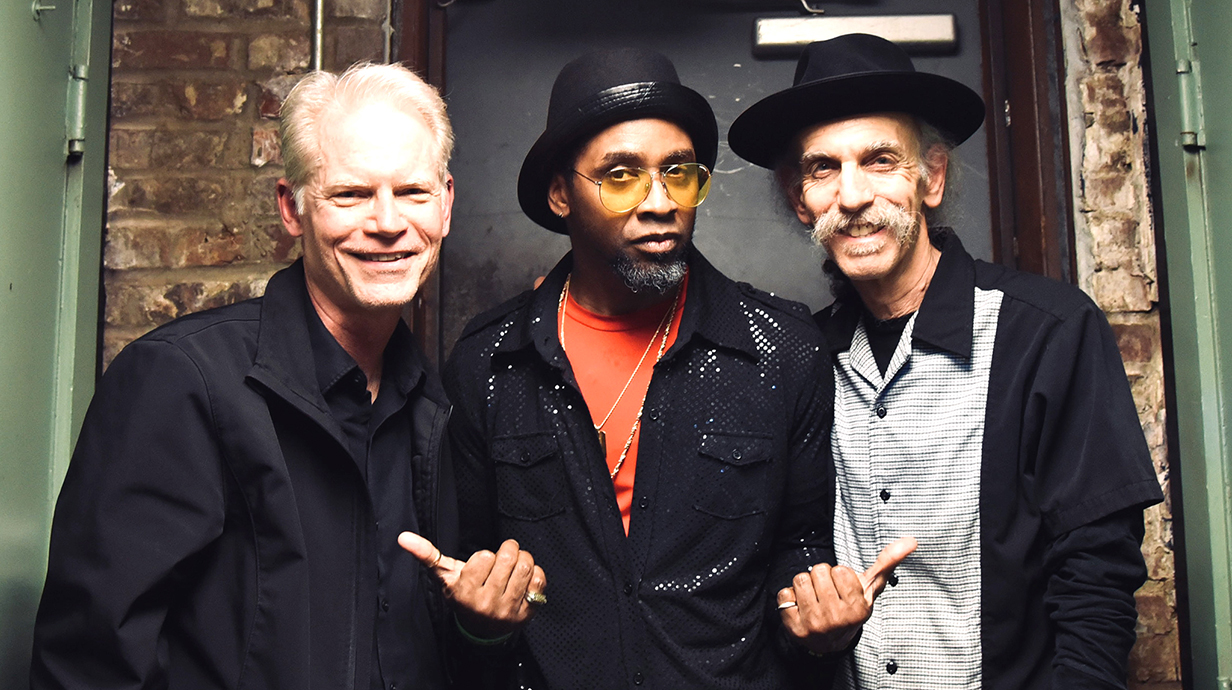 Three men pose for a photo, with the one in the center pointing at the other two.