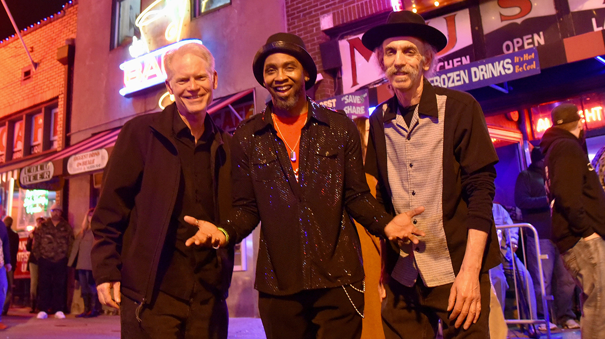 Three men stand together on a brightly lit street at night.