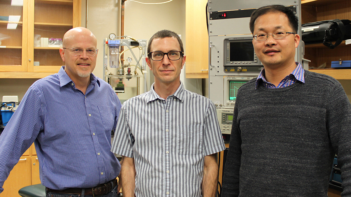 Three men stand in a physics laboratory.