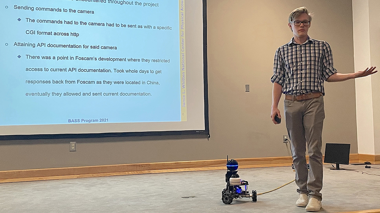 A young man gestures in front of a projection screen with a small robotic vehicle at his feet.