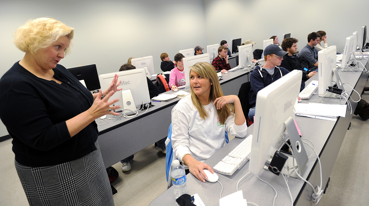 An older woman talks with a young woman seated at a computer in a classroom filled with students at computers.