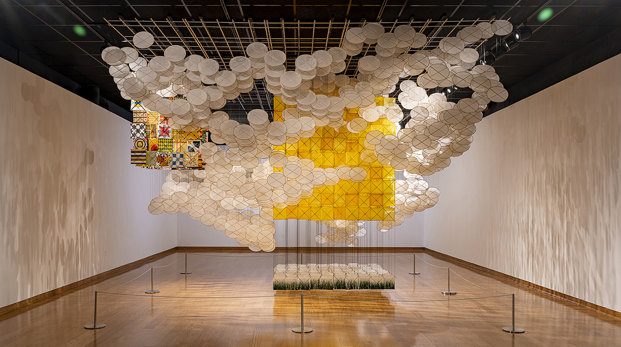 A large piece of three-dimensional artwork made of paper and string, hangs in a museum exhibit.