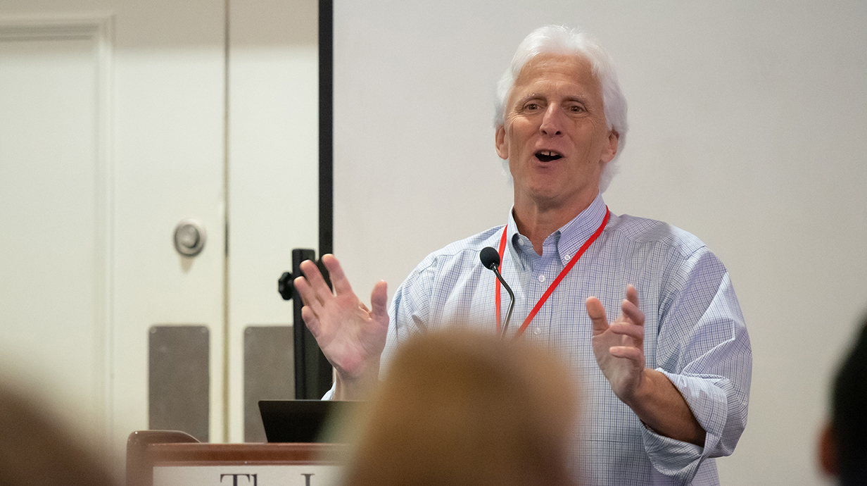 A white-haired man talks to a crowd from a podium.