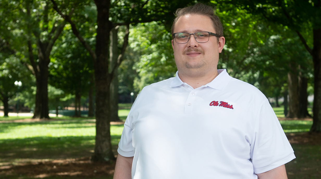 A man wearing a white Ole Miss polo shirt stands in a tree-shaded park.