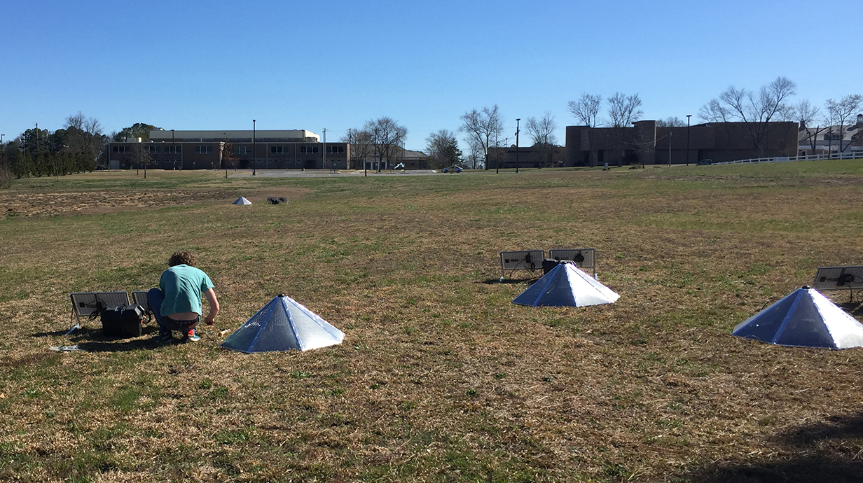 A young man kneels to work on solar panels and other equipment near three silver, cone-shaped objects in a field.