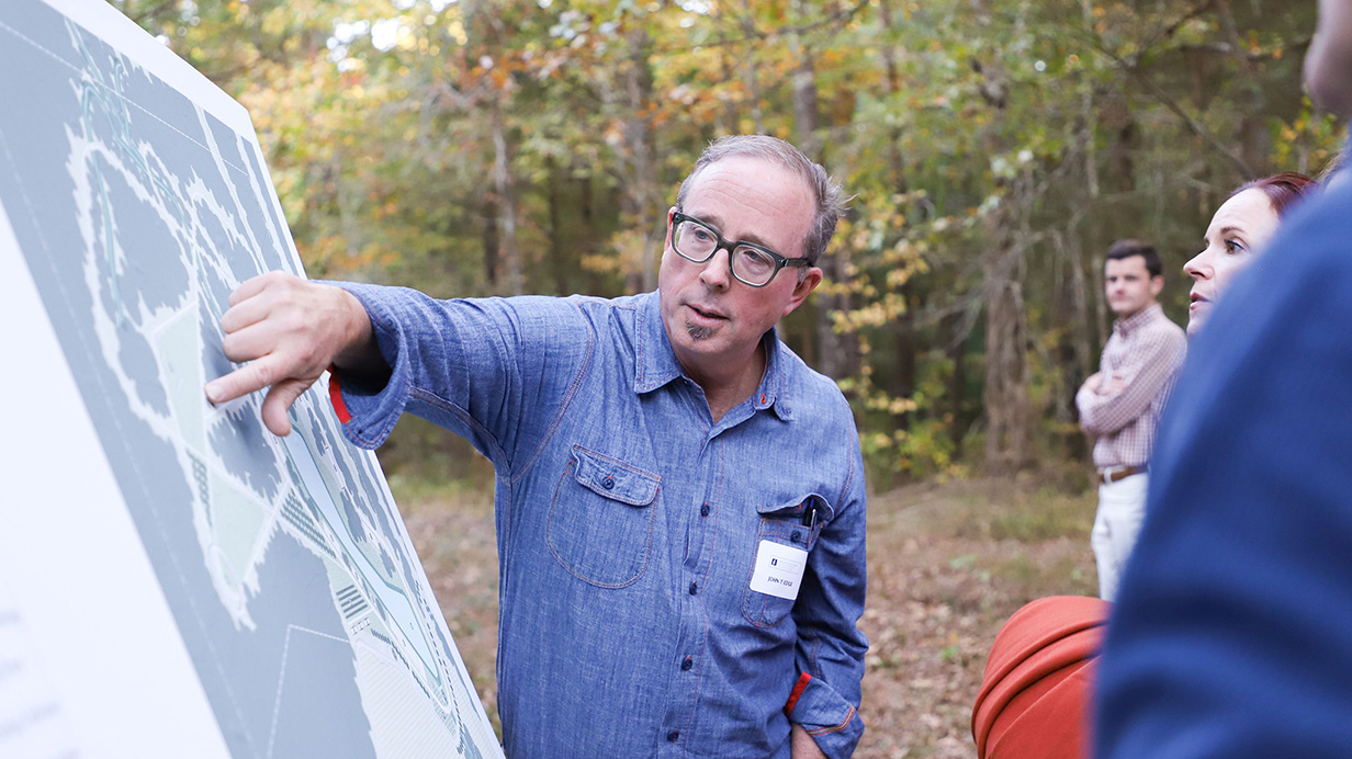 A man points to a spot on a large mounted map as other people standing in a clearing look on.