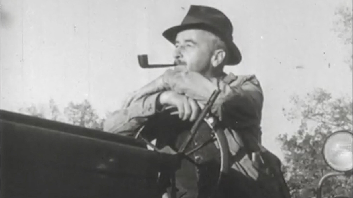 Black-and-white photo of a man wearing a hat and smoking a pipe as he sits on a tractor.