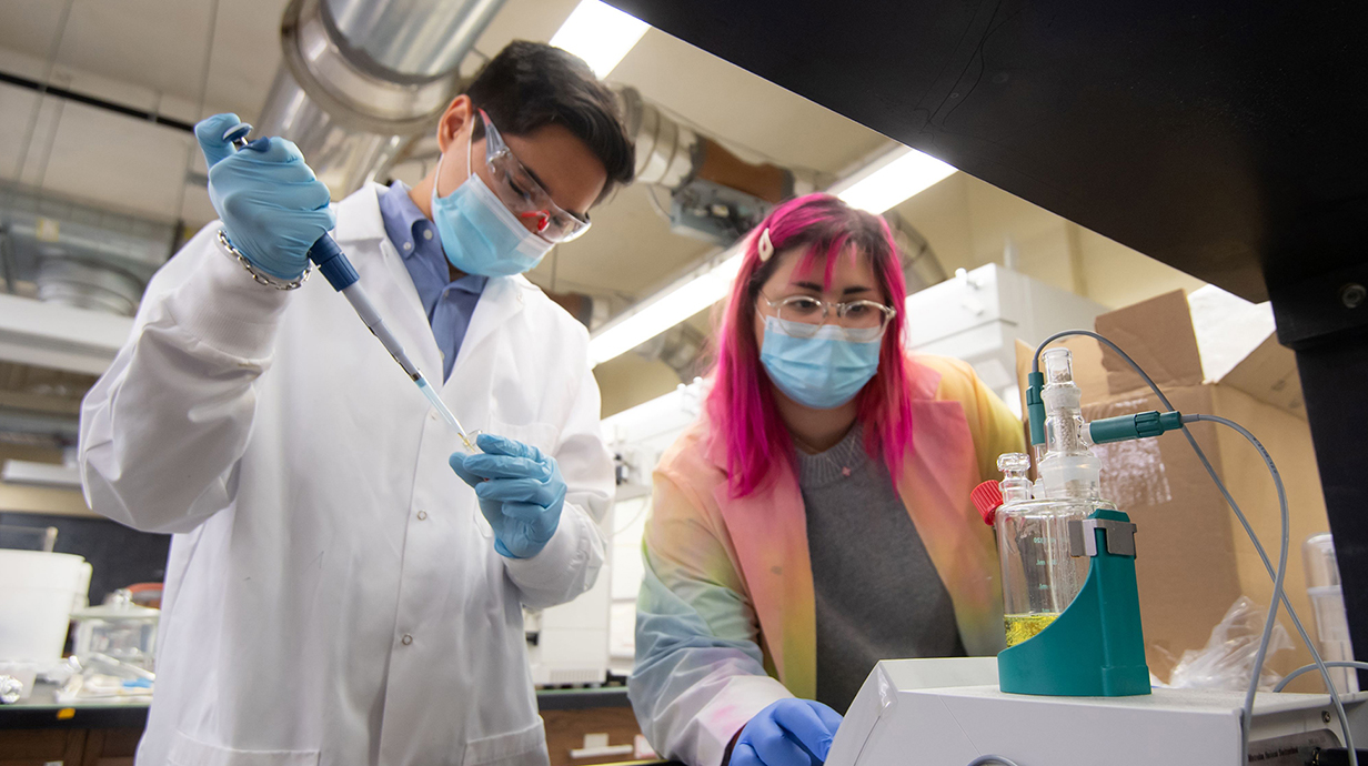 A woman and a man, both wearing white lab coats, safety glasses and blue gloves, work with a pipette and beaker of liquid in a laboratory.
