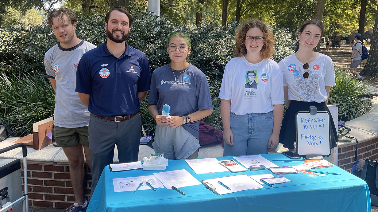 Two young men and three young women stand behind an outdoor table covered with leaflets and forms and a sign reading 'Register to Vote!'