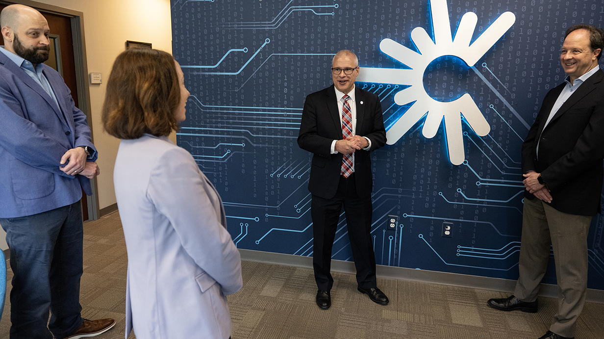 Three men and a woman talk in front of a high-tech corporate logo.
