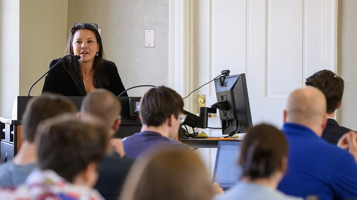 A woman speaks into a microphone at the front of a classroom full of college students.