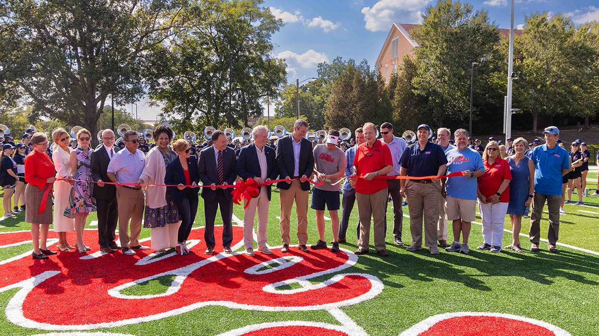 A large group of people participate in a ribbon-cutting ceremony on a band practice field.