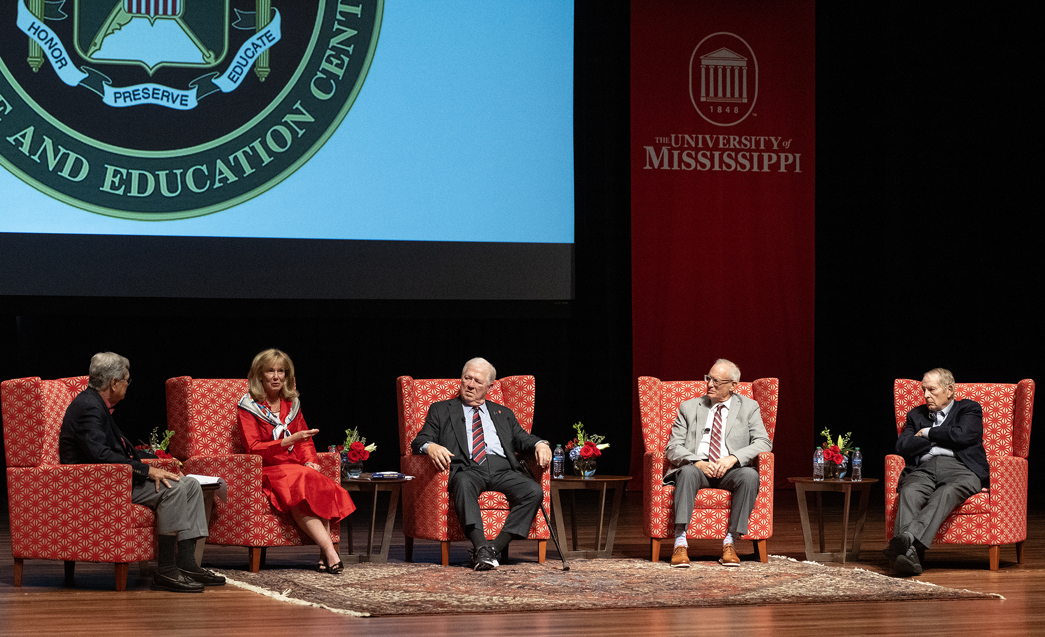 Four people sit in red chairs on a stage discussing a topic. 