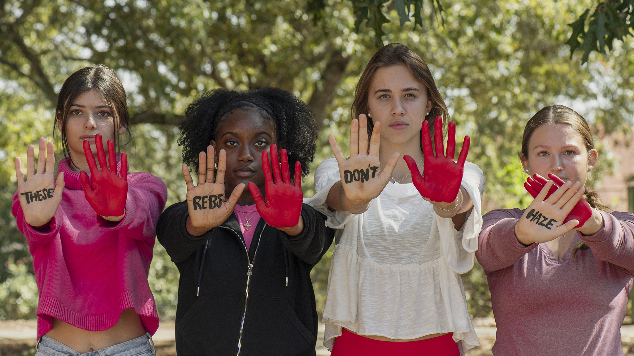 Four students look straight ahead with their palms out. Each has one palm painted red. The other palm has a word on it. The words spell These Rebs Don't Haze