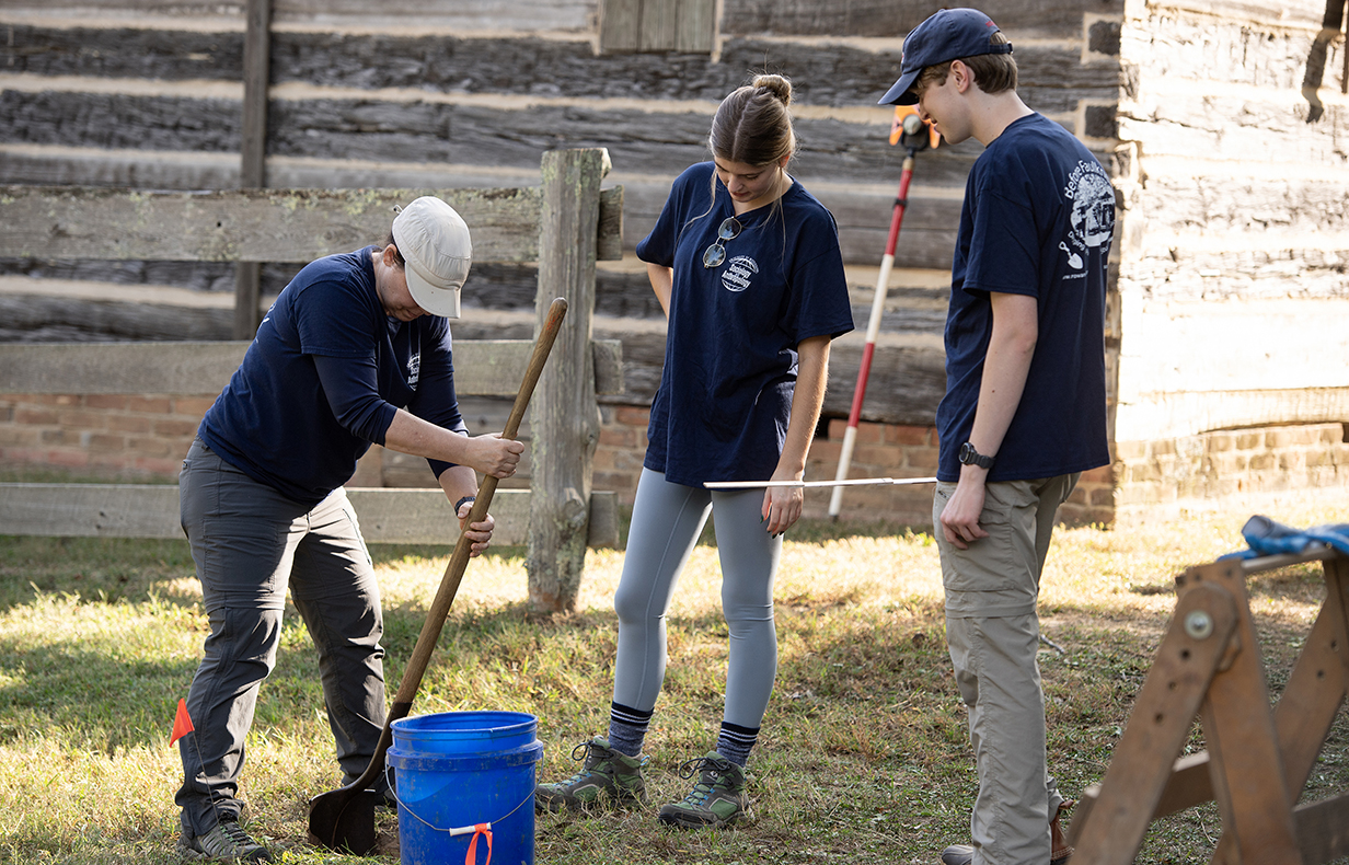 A woman uses a shovel to dig a hole while a young woman and man watch.