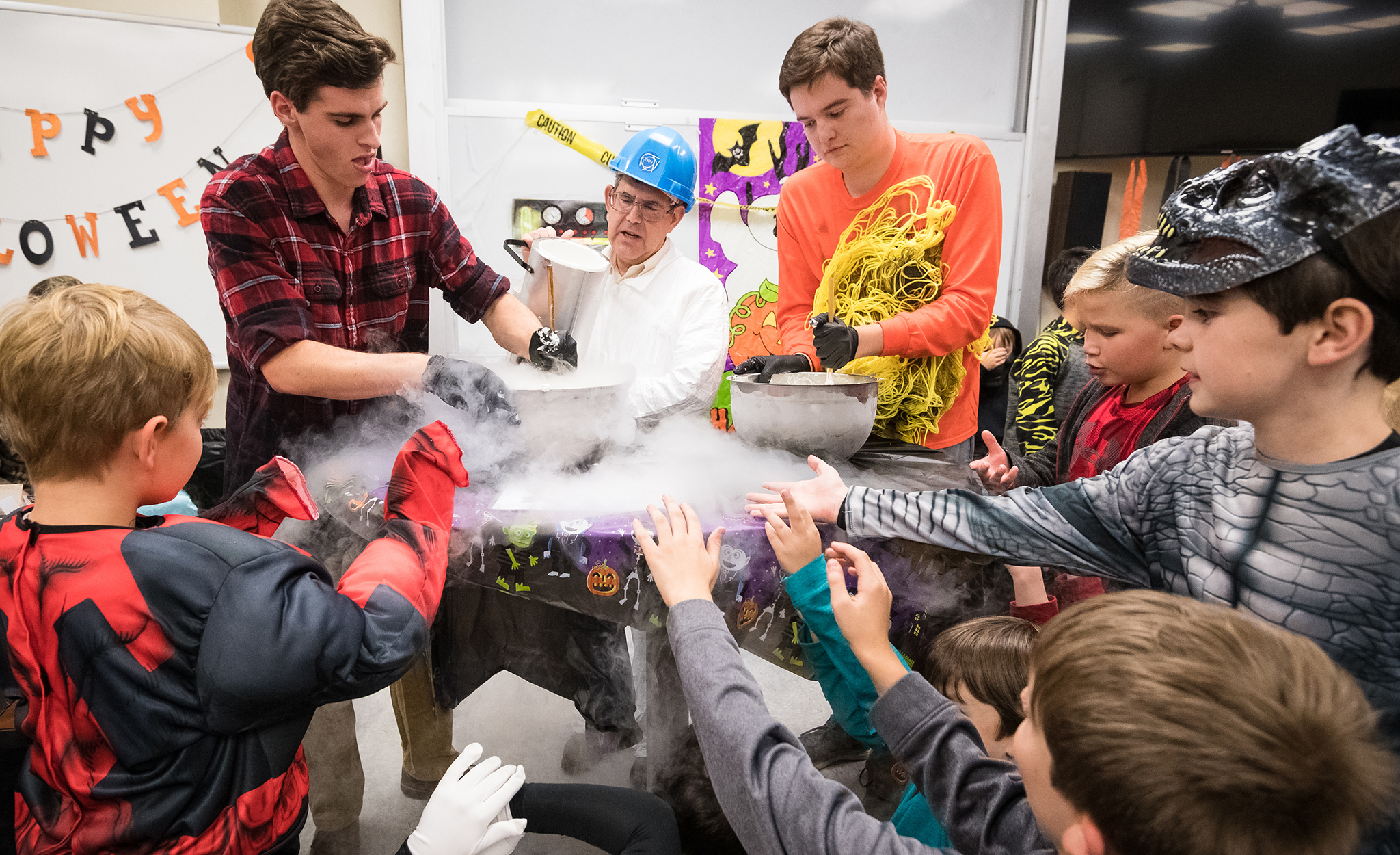 University professors and young students gather around a table with a mist coming off. Halloween decorations hang in the background. 