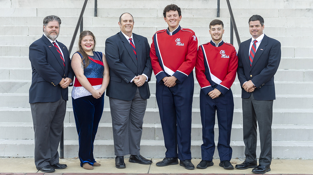 Three men and three young people, wearing band uniforms, pose for a photo in front of concrete steps.