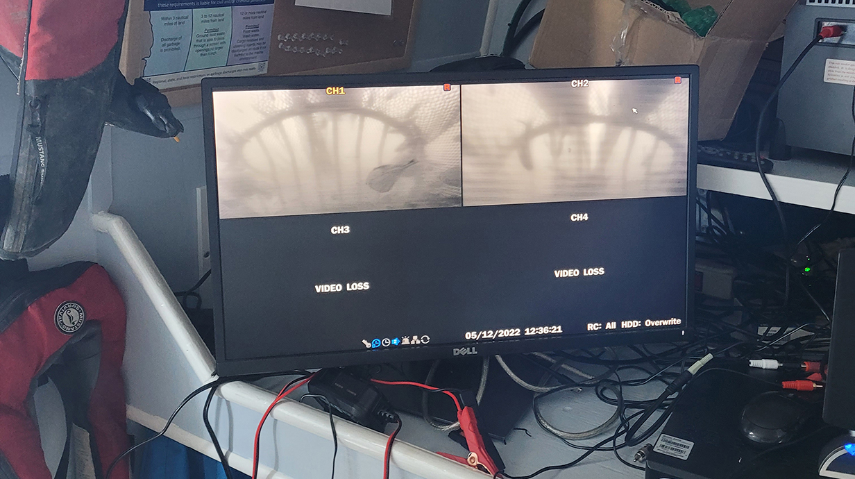 A video screen sitting amongst a pile of wires and equipment shows images of underwater nets.