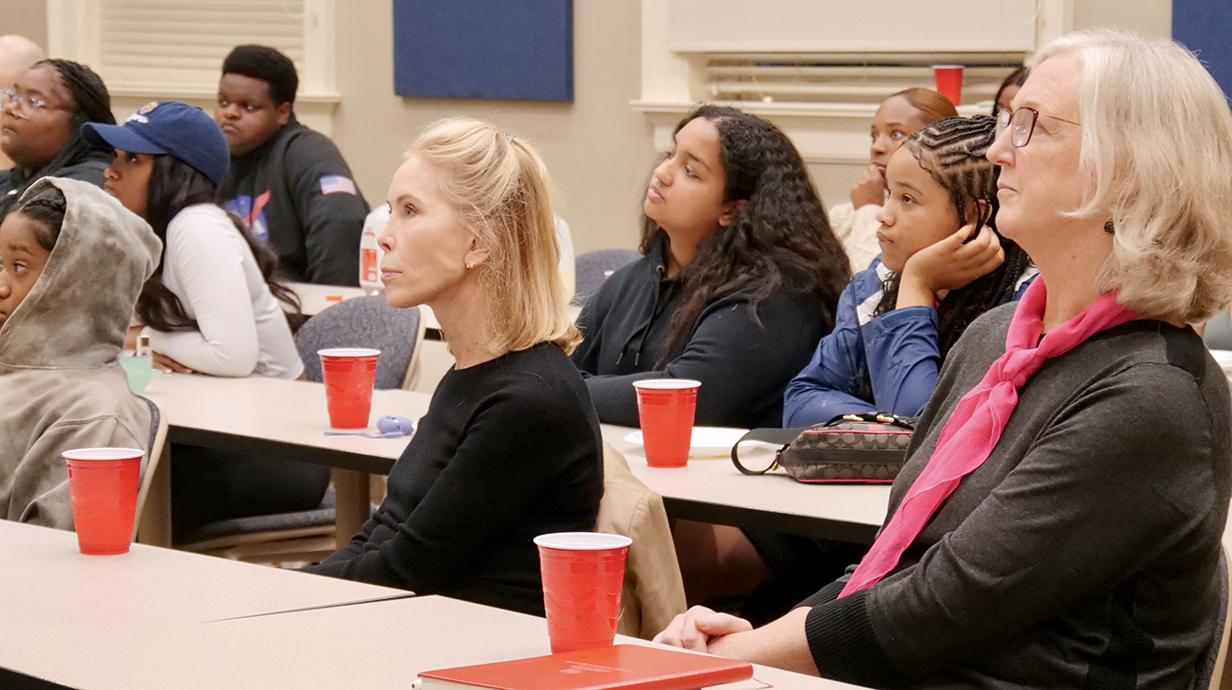 A woman sits with students and administrators in a classroom.