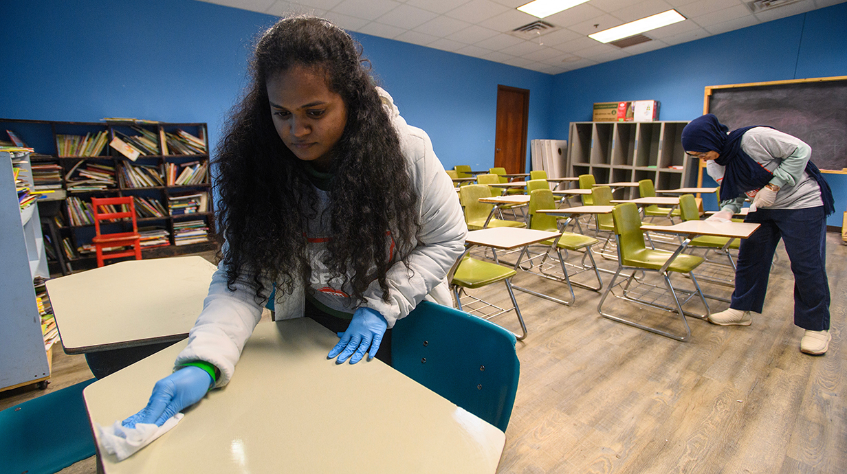 Two women wipe down desks in a classroom.