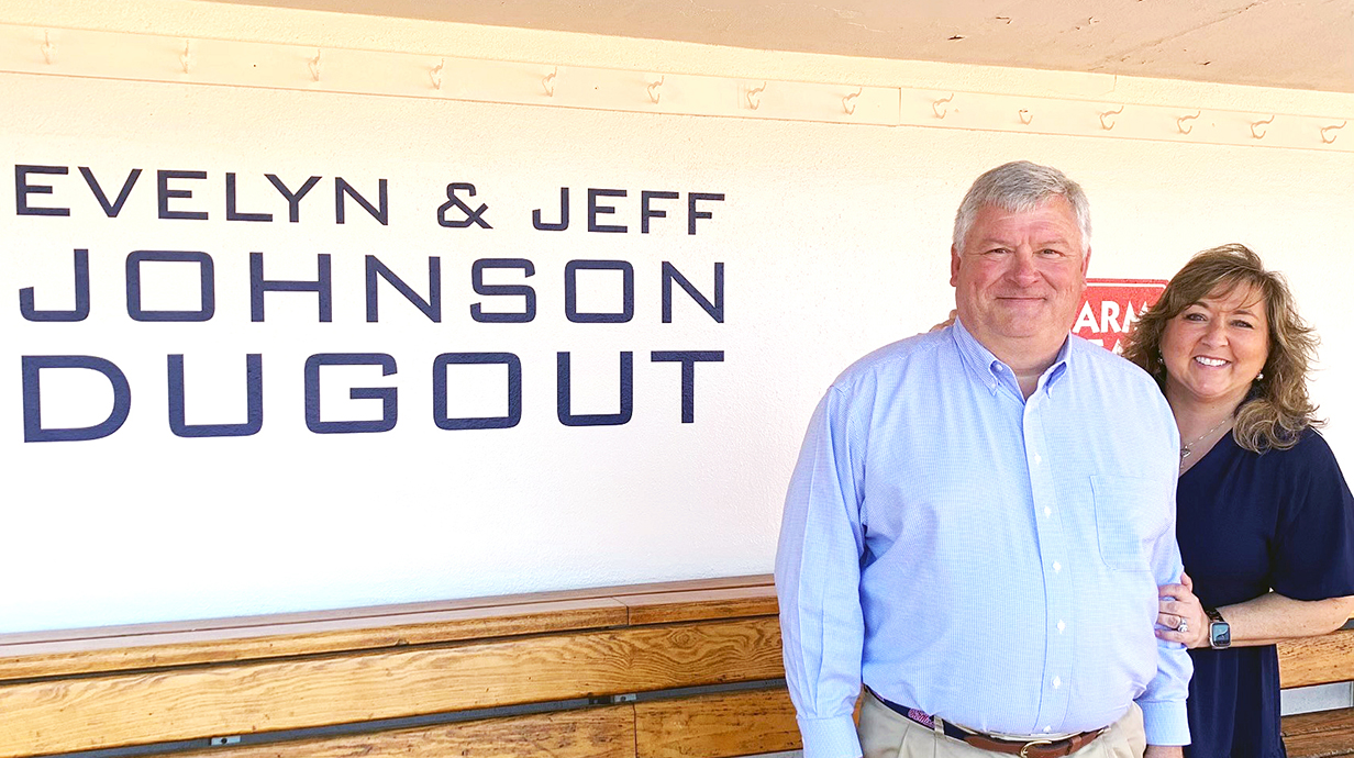 A man and a woman stand in front of a sign reading 'Evelyn & Jeff Johnson Dugout'