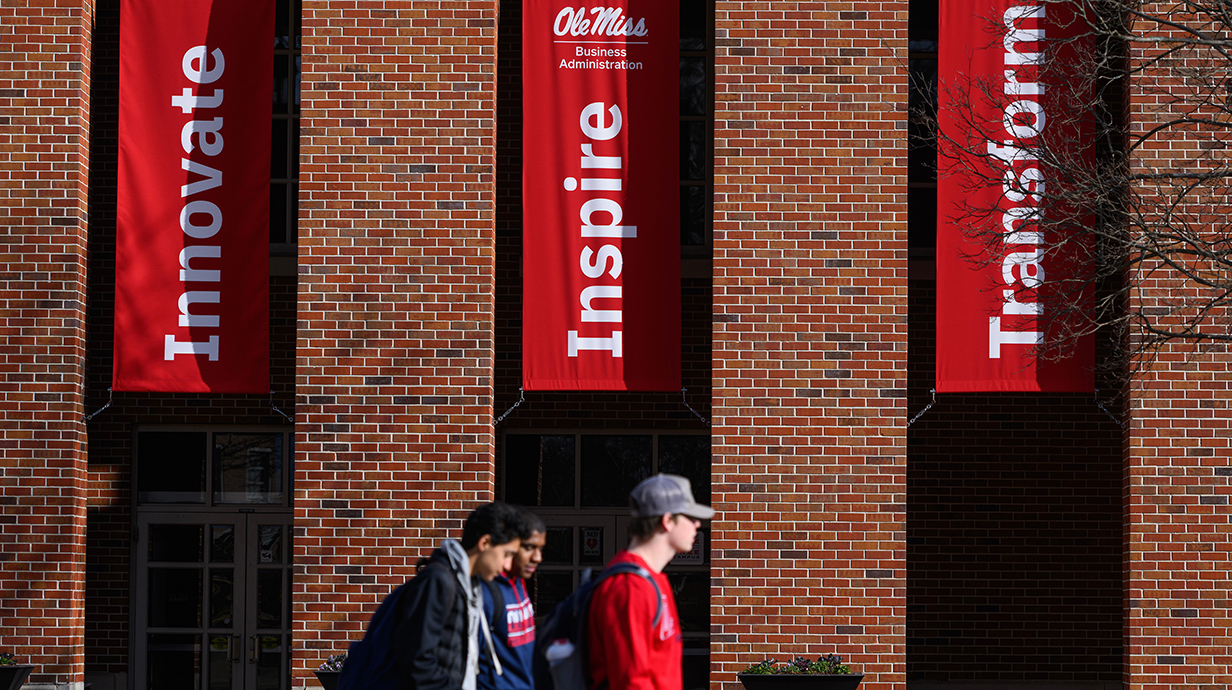 Three young men walk in front of a brick building that displays banners reading Innovate, Inspire and Transform.