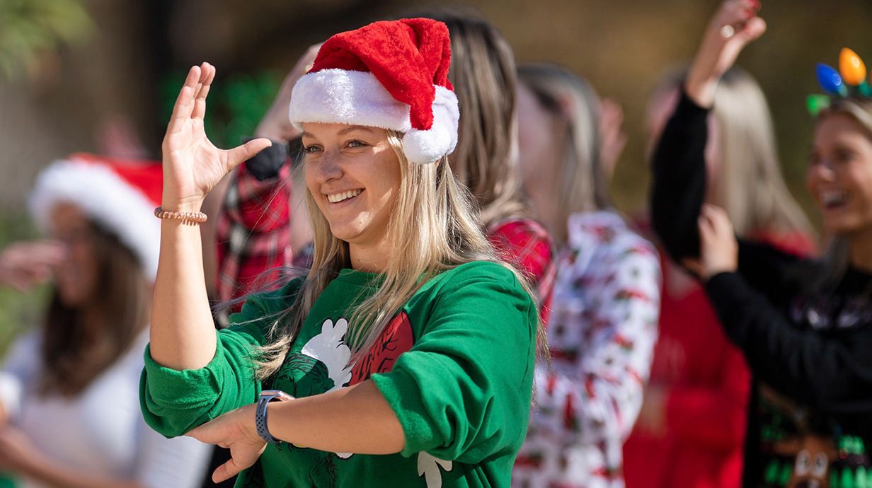 Several young women wearing Christmas-themed clothing perform American Sign Language.