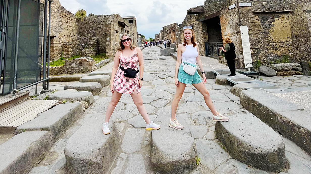 Two young women cross a stone street in the Pompei archaeological site.