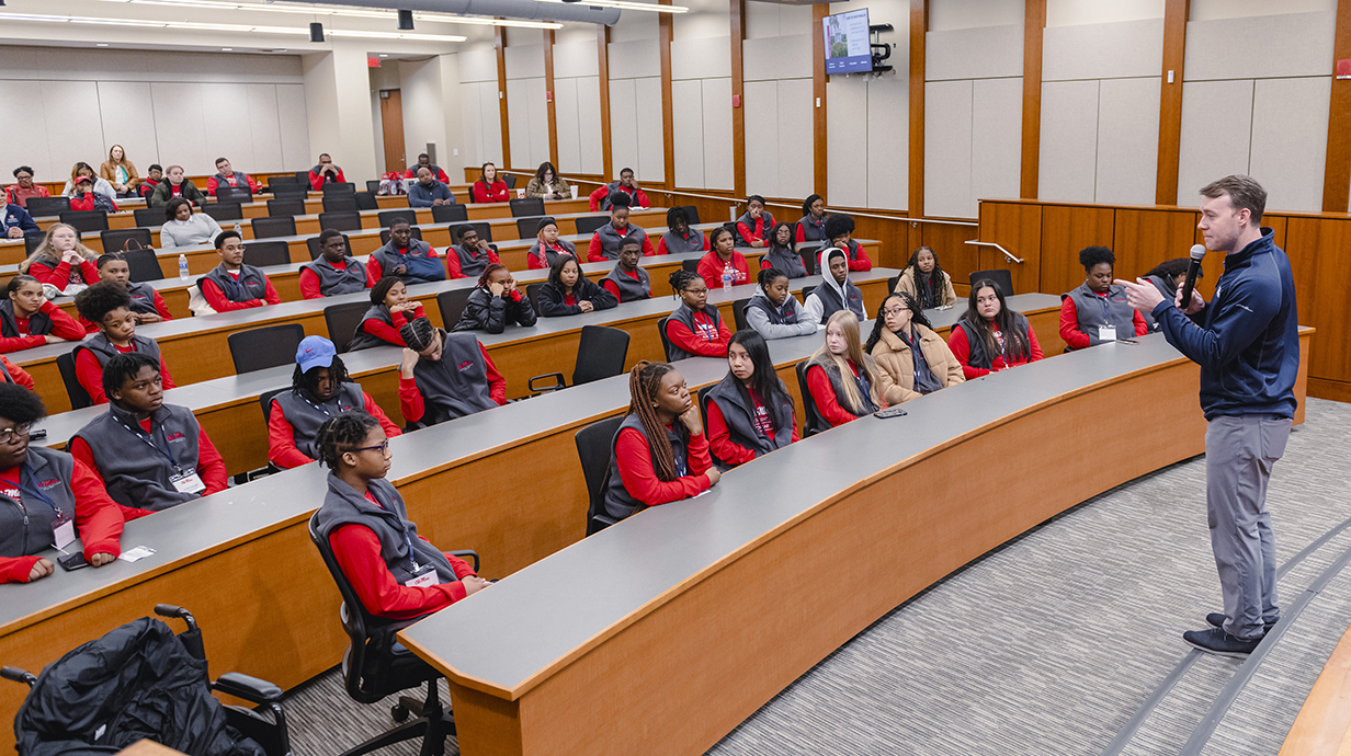 A man with a microphone speaks to high school students in a tiered auditorium.