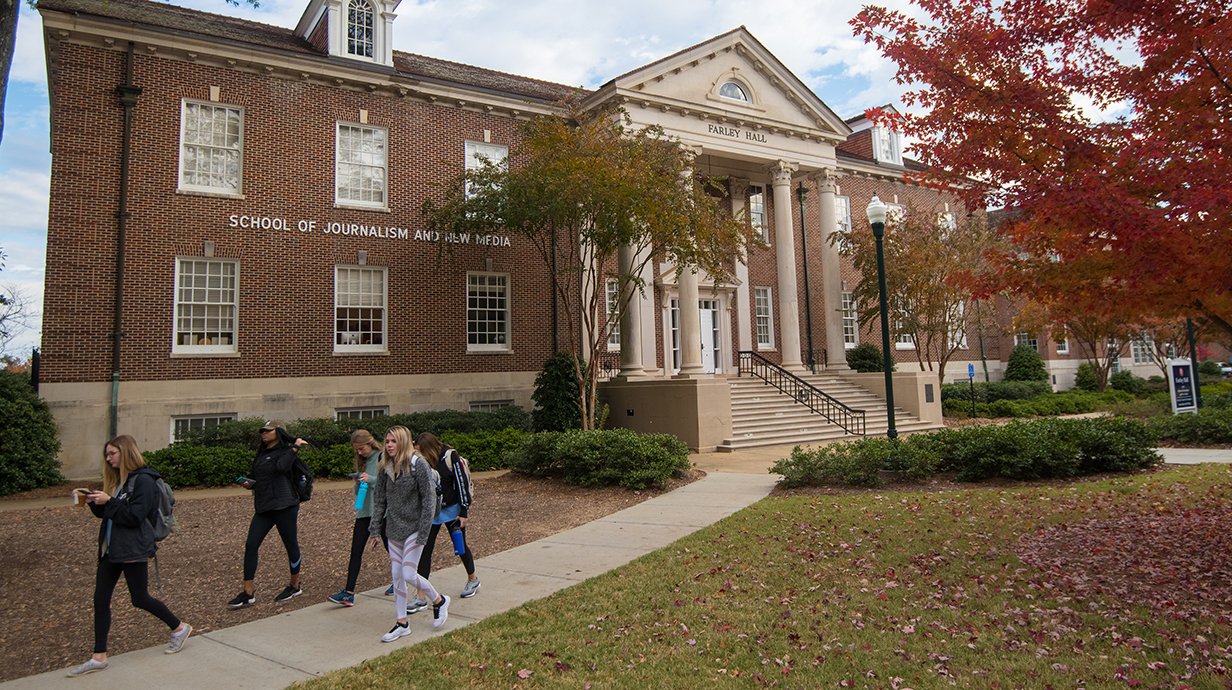 A group of students walk in front of a building with a sign reading 'School of Journalism and New Media'