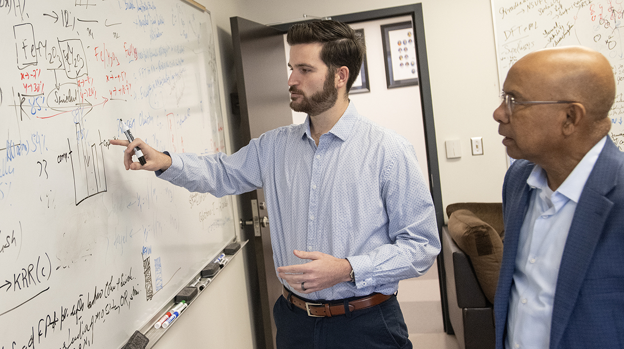 A young man points out equations on a whiteboard to an older man.