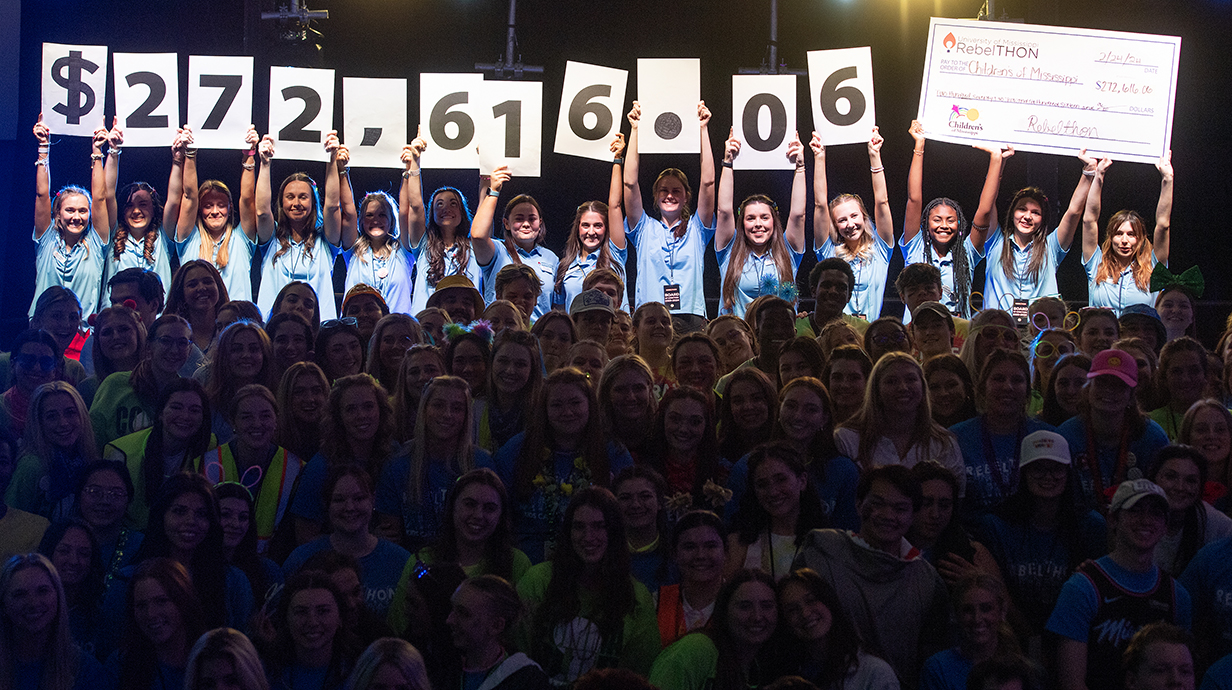 A group of young women hold up signs spelling out "$272,616.06" and a giant check while a crowd of young people pose in front.