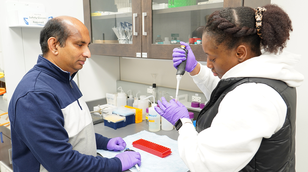 A young woman uses a pipette to transfer a liquid in a laboratory while a man watches.