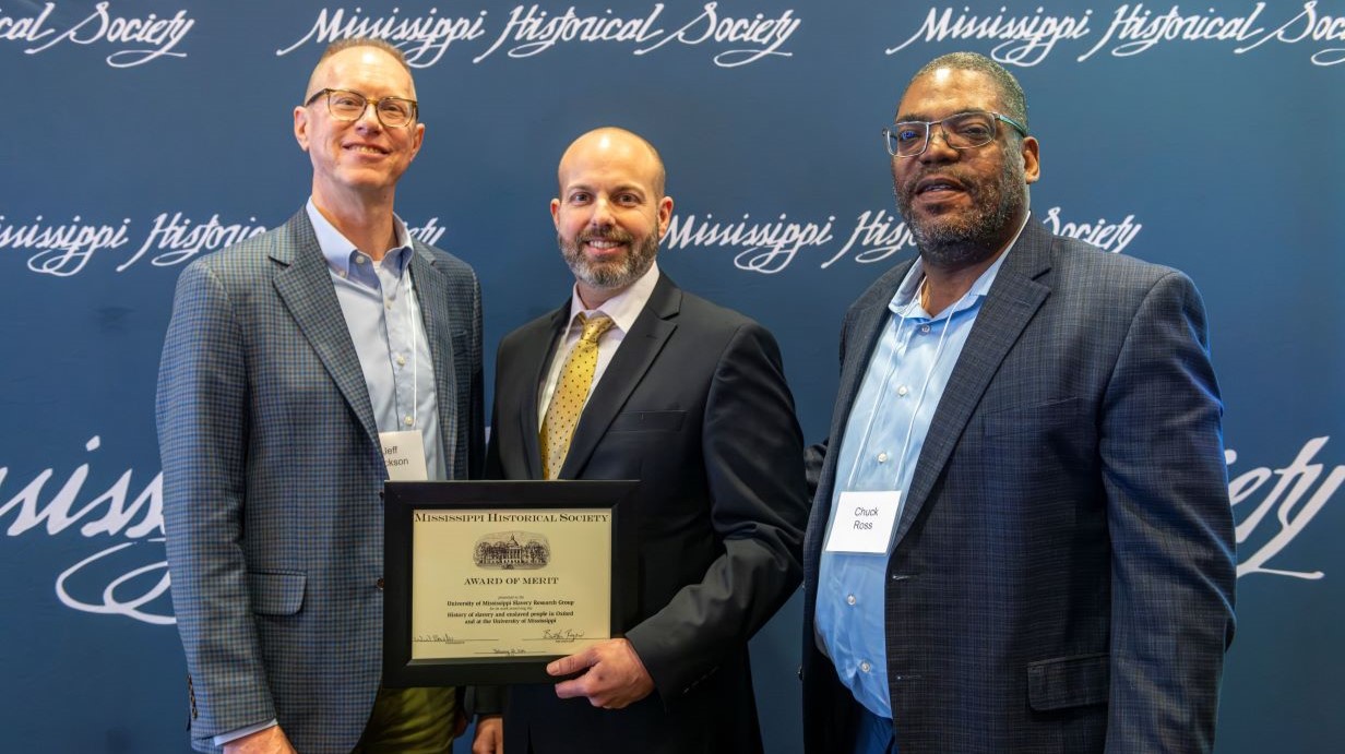 Three men stand in a row in front of a blue background, the middle one holding an award certificate.