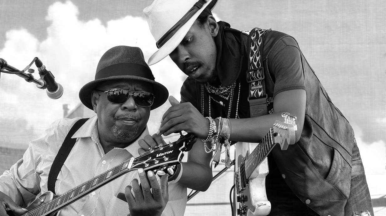 Two men adjust a guitar during an onstage performance.