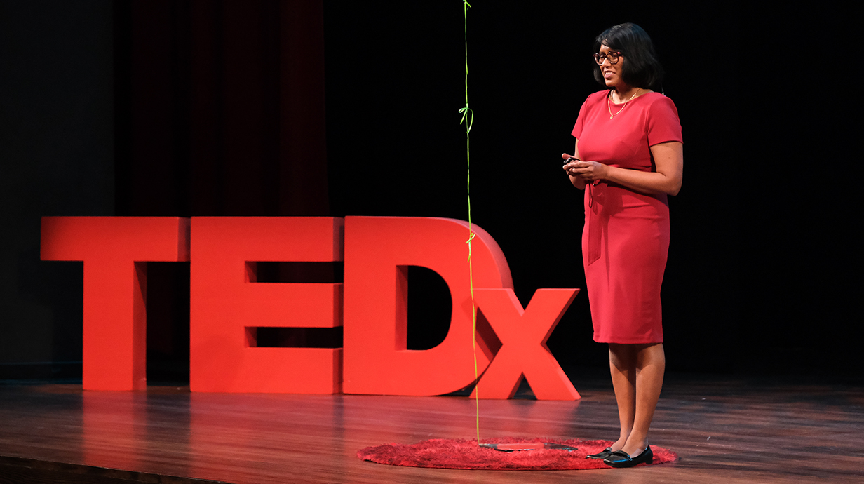 A woman speaks on a stage next to a red TEDx logo.