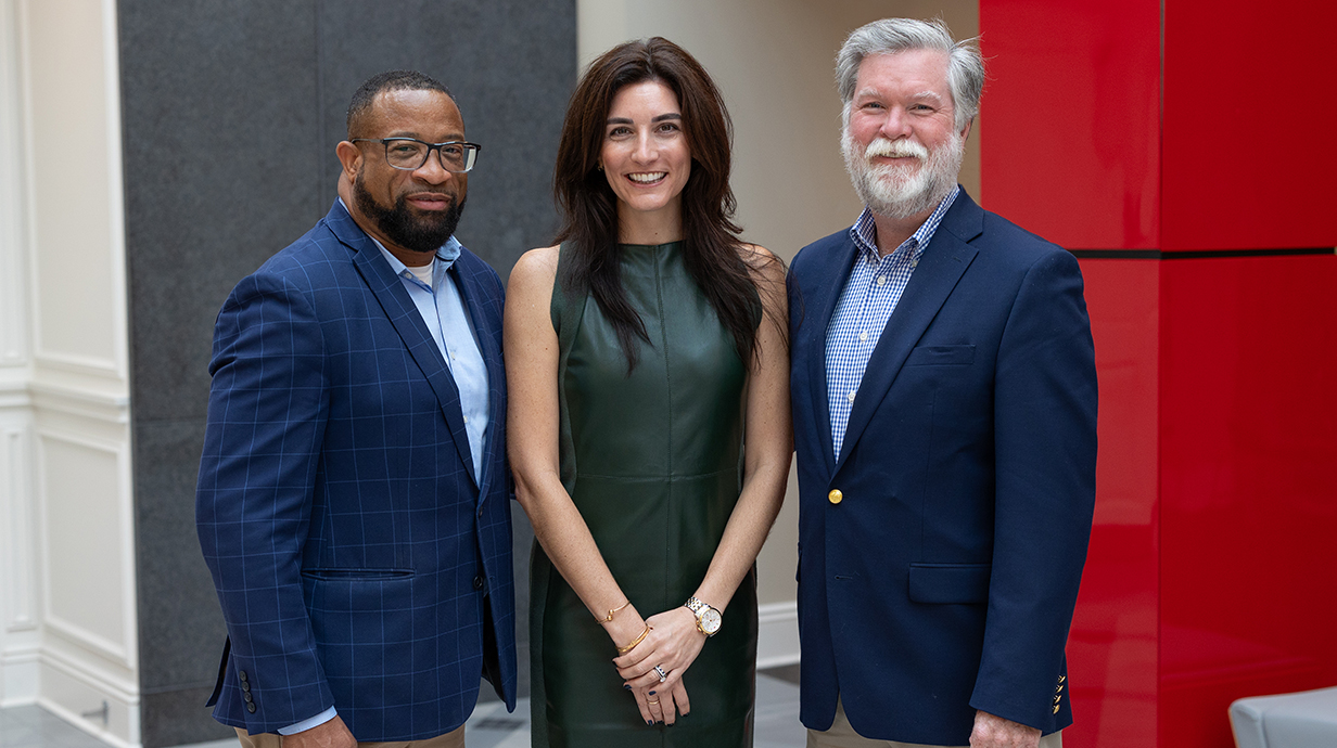 Two men and a woman in business clothes stand in front of a red and gray background.